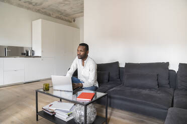 Nachdenklicher Mann sitzt auf der Couch in einer modernen Wohnung und benutzt einen Laptop - AHSF02722
