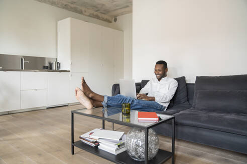 Porträt eines lächelnden Mannes, der auf einer Couch in einer modernen Wohnung sitzt und einen Laptop benutzt - AHSF02725