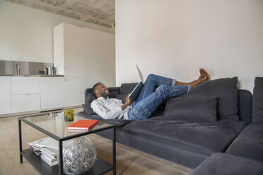 Smiling man lying on couch in modern apartment using laptop - AHSF02726