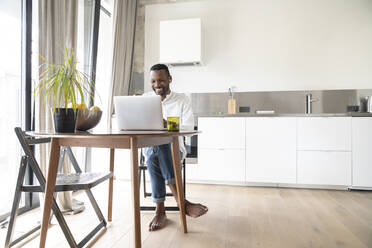 Porträt eines lächelnden Mannes, der an einem Tisch in einer modernen Wohnung sitzt und einen Laptop und Kopfhörer benutzt - AHSF02727