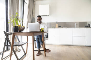 Portrait of smiling man sitting at table in modern apartment using laptop and earphones - AHSF02727