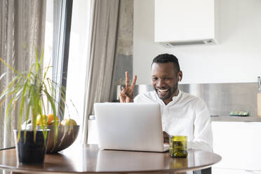 Portrait of happy man sitting at kitchen table during video chat showing victory sign - AHSF02728