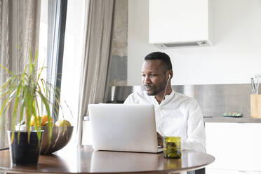 Portrait of man using laptop and earphones at home - AHSF02729