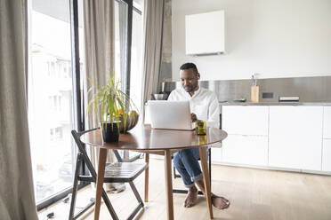 Porträt eines Mannes, der in einer modernen Wohnung am Tisch sitzt und einen Laptop und Kopfhörer benutzt - AHSF02730