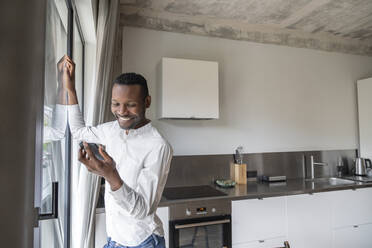 Porträt eines lächelnden Mannes, der in seiner Küche an der Balkontür steht und auf sein Mobiltelefon schaut - AHSF02734