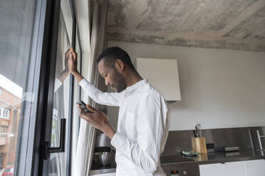 Pensive man standing at French door at home with mobile phone - AHSF02735