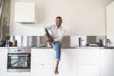 Portrait of smiling man sitting barefoot on kitchen counter at home - AHSF02744