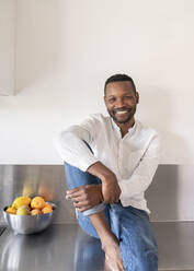Porträt eines lächelnden Mannes, der zu Hause auf dem Küchentisch sitzt - AHSF02745