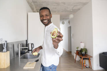 Portrait of smiling man with sliced avocado in his kitchen - AHSF02751
