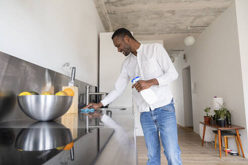 Smiling man standing in kitchen of his apartment cleaning countertop - AHSF02755