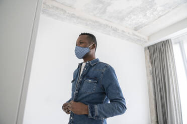 Man wearing reusable face mask indoors putting on his jeans jacket - AHSF02763