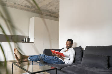 Lächelnder Mann sitzt auf der Couch in einer modernen Wohnung und liest ein Buch - AHSF02766