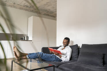 Smiling man sitting on couch in modern apartment reading a book - AHSF02766