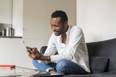 Lächelnder Mann sitzt zu Hause auf der Couch und benutzt sein Smartphone - AHSF02767