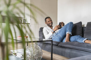Porträt eines lächelnden Mannes, der in einer modernen Wohnung auf der Couch sitzt und ein Smartphone benutzt - AHSF02768