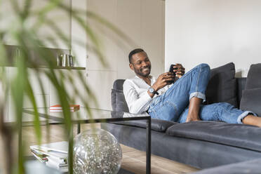 Portrait of smiling man sitting on couch in modern apartment using smartphone - AHSF02768