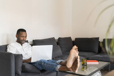 Portrait of man sitting on couch with feet up using laptop - AHSF02772