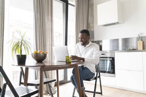 Lächelnder Mann, der zu Hause am Tisch sitzt und einen Laptop benutzt, lizenzfreies Stockfoto