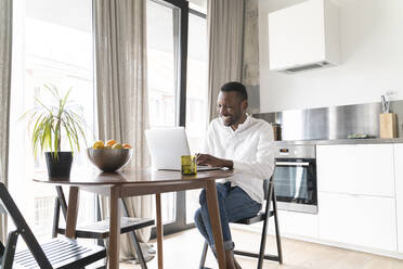 Smiling man sitting at table at home using laptop - AHSF02773