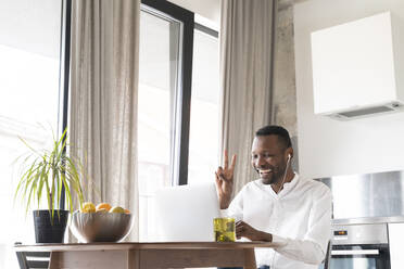 Portrait of happy man sitting at kitchen table during video chat showing victory sign - AHSF02774