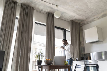 Man standing at Frencch door in modern apartment using smartphone - AHSF02775