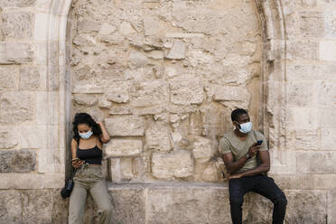 Man and woman wearing masks while using smart phones against wall - EGAF00176