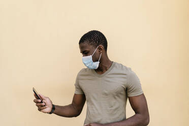 Mann trägt eine Maske und benutzt ein Smartphone an einer beigefarbenen Wand - EGAF00180