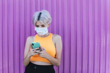 Blonde Frau mit Gesichtsmaske, die ein Smartphone vor einer lila Wand benutzt - TCEF00739