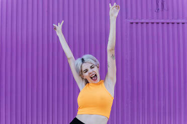 Beautiful blond woman standing in front of purple wall, raising arms, laughing - TCEF00743