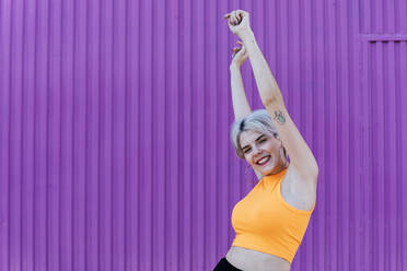 Beautiful blond woman standing in front of purple wall, raising arms, laughing - TCEF00744