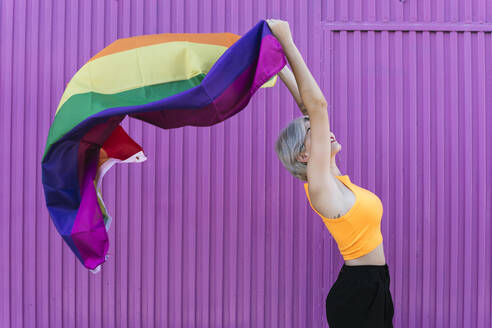 Smiling blond woman holding LGBT rainbow flag in front of purple wall - TCEF00745