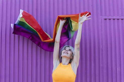 Lächelnde blonde Frau hält LGBT-Regenbogenfahne vor lila Wand - TCEF00746