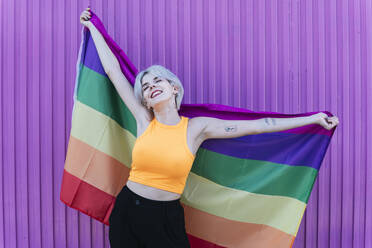 Smiling blond woman holding LGBT rainbow flag in front of purple wall - TCEF00747