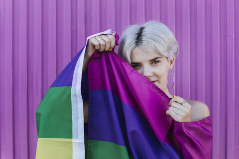 Lächelnde blonde Frau, eingewickelt in eine LGBT-Regenbogenfahne vor einer lila Wand, lizenzfreies Stockfoto