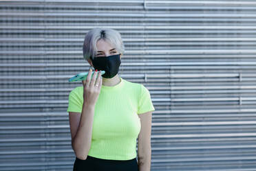 Blonde Frau steht vor einem Metallhintergrund, trägt eine Gesichtsmaske und benutzt ein Smartphone - TCEF00752