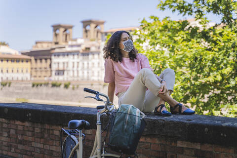 Woman wearing protective mask while looking away and sitting on retaining wall by bicycle in city during coronavirus stock photo