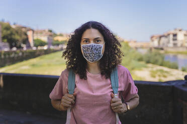 Woman wearing protective face mask standing with backpack in city against sky - FMOF00993