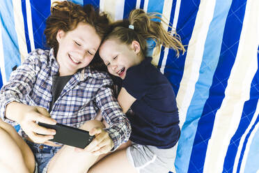 Glückliche Mädchen teilen sich ein Smartphone, während sie auf einer Picknickdecke liegen - IHF00351