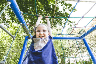 Lächelndes süßes Mädchen hängt am Klettergerüst auf einem Spielplatz im Park - IHF00353