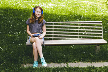 Smiling girl listening music through headphones while sitting on bench at park - IHF00356