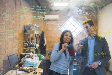 Businessman and woman brainstorming taking notes on glass pane in office - MEUF00848