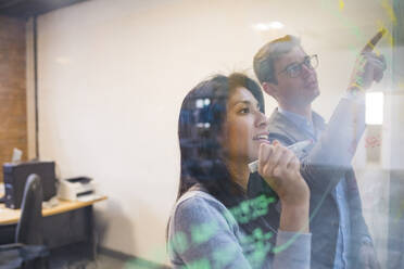 Businessman and woman brainstorming taking notes on glass pane in office - MEUF00849