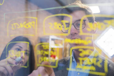 Businessman and woman brainstorming taking notes on glass pane in office - MEUF00850