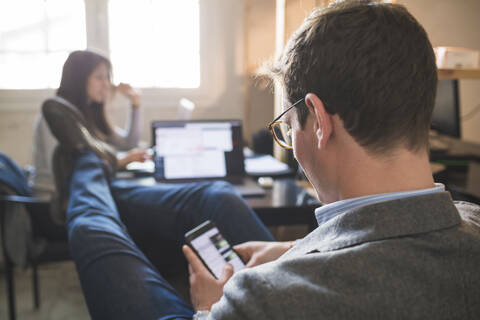 Entspannter Geschäftsmann, der sein Smartphone am Schreibtisch benutzt, während eine Frau im Hintergrund arbeitet, lizenzfreies Stockfoto
