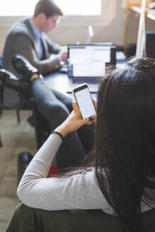 Entspannte Frau mit Smartphone am Schreibtisch mit Geschäftsmann im Hintergrund arbeiten - MEUF00871