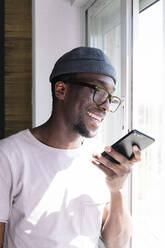 Happy young man using smart phone by window - JCMF00829