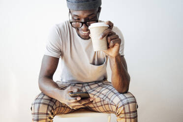 Lächelnder Mann, der eine Textnachricht schreibt, während er eine Kaffeetasse hält und vor einem weißen Hintergrund sitzt - JCMF00830