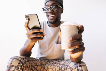Smiling young man using smart phone while sitting with disposable coffee cup against white background - JCMF00832