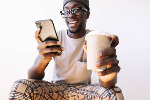 Lächelnder junger Mann, der sein Smartphone benutzt, während er mit einer Einweg-Kaffeetasse vor weißem Hintergrund sitzt - JCMF00832