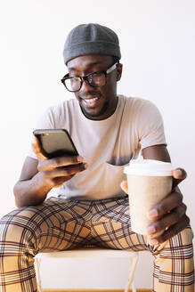 Junger Mann, der ein Smartphone benutzt, während er mit einer Kaffeetasse vor einem weißen Hintergrund sitzt - JCMF00833